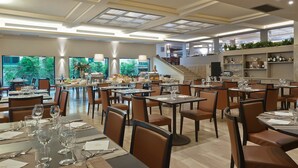 A spacious dining area with multiple tables and chairs, large windows with a view of greenery, and a staircase leading to an upper level.