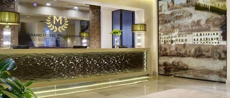 A Grand Hotel Mediterraneo reception area with a modern design, including a reception desk, a decorative wall, and a large painting of a landscape.