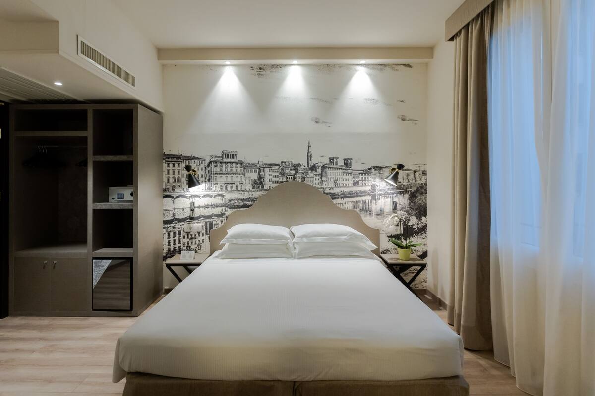 Une chambre d’hôtel avec un grand lit, une armoire, une table de chevet avec une lampe et une fresque murale représentant un paysage urbain.