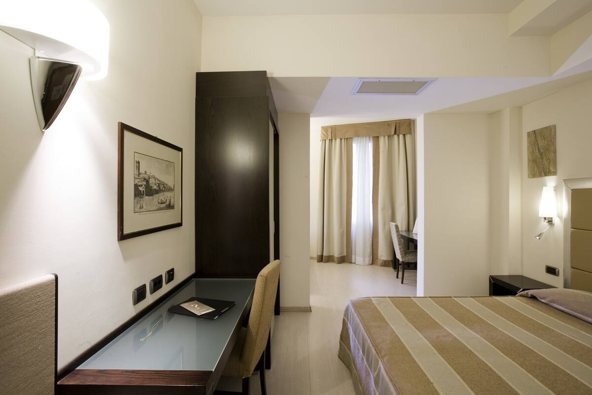 Une chambre d’hôtel avec un lit, un bureau, une chaise, un tableau encadré au mur et une fenêtre avec des rideaux.