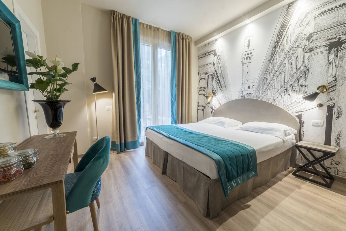 Une chambre d’hôtel avec un lit, un bureau et une chaise, une plante en pot et un mur orné d’une fresque représentant un paysage urbain.