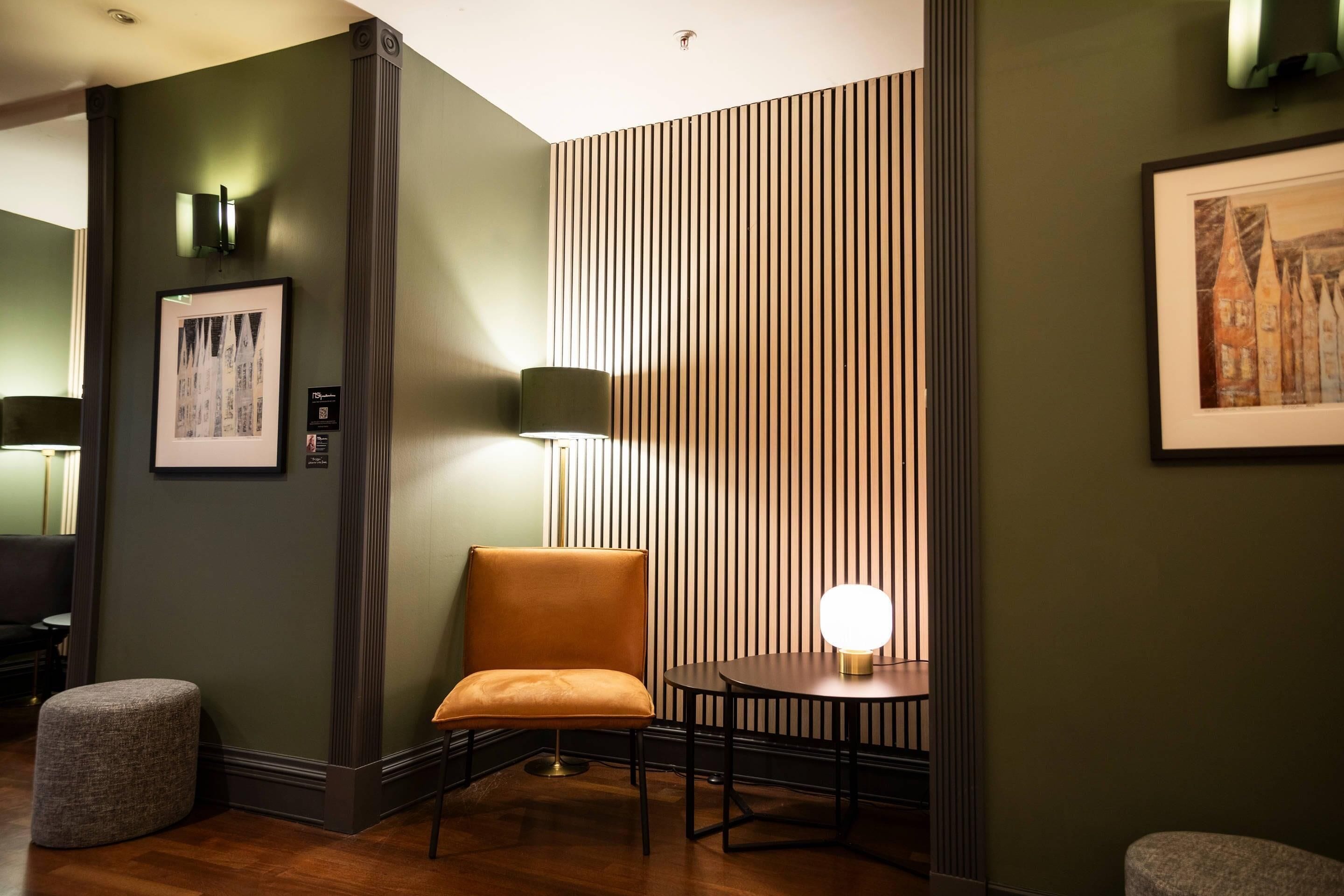 A room with a green wall, a framed picture, a tall lamp, a round table with a light, and a cushioned chair.