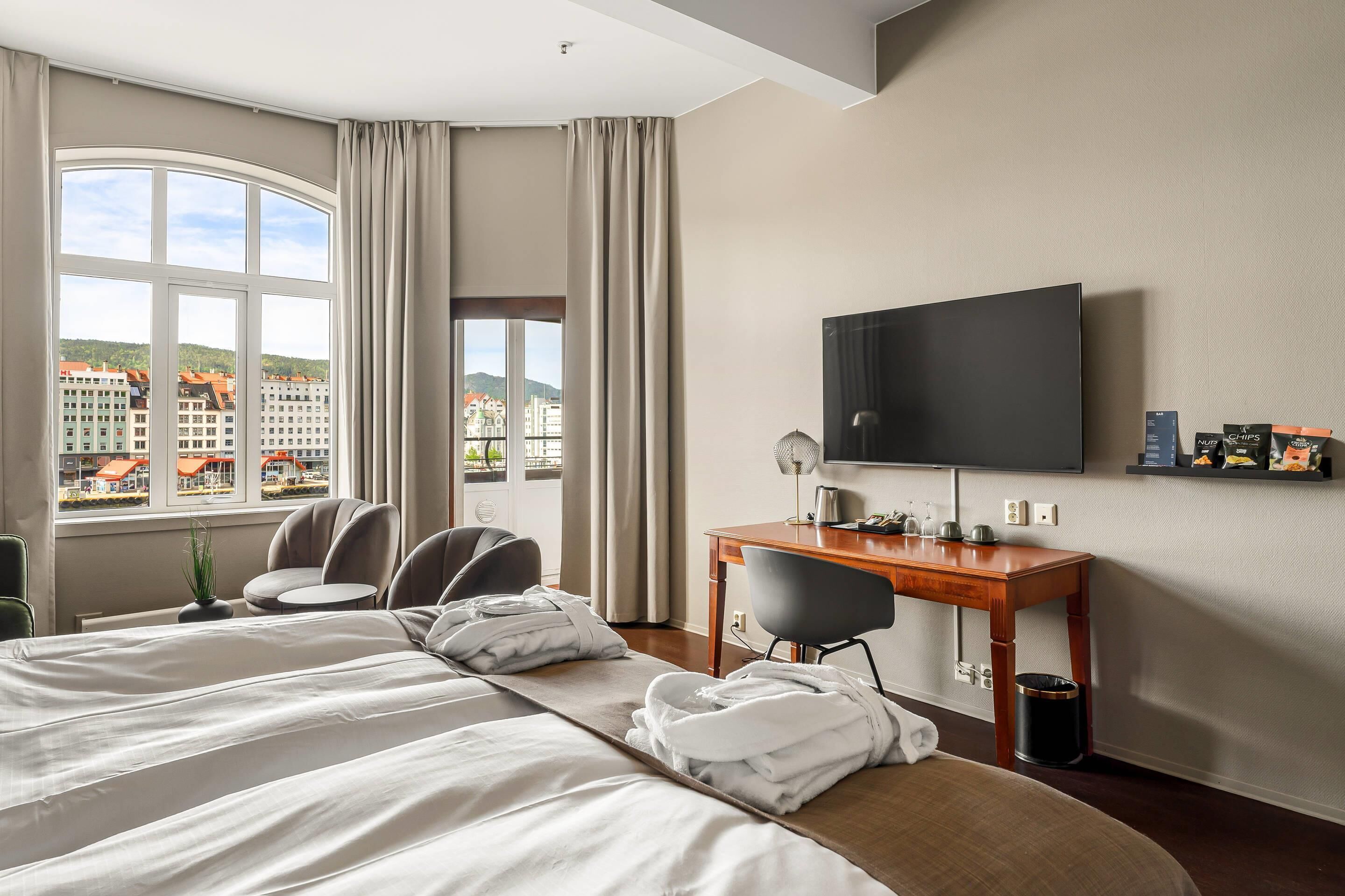 A hotel room with a large bed, a desk, a television, and a view of buildings outside the window.