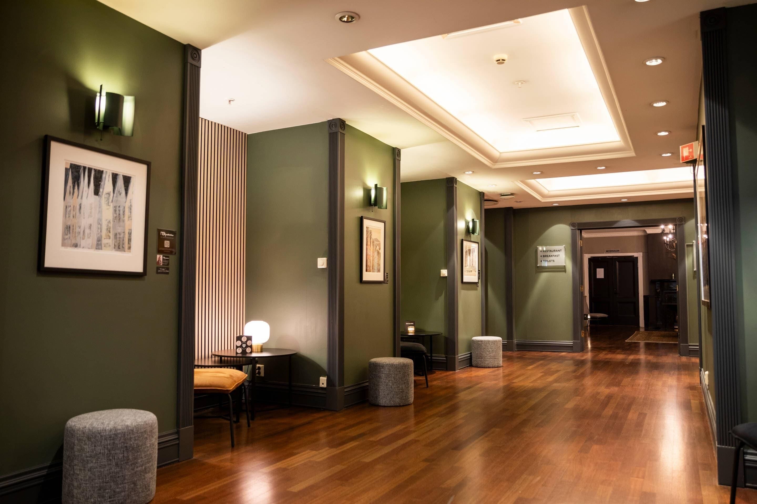 A hotel lobby with a wooden floor, green walls, and framed artwork. There are seating areas with tables and chairs.