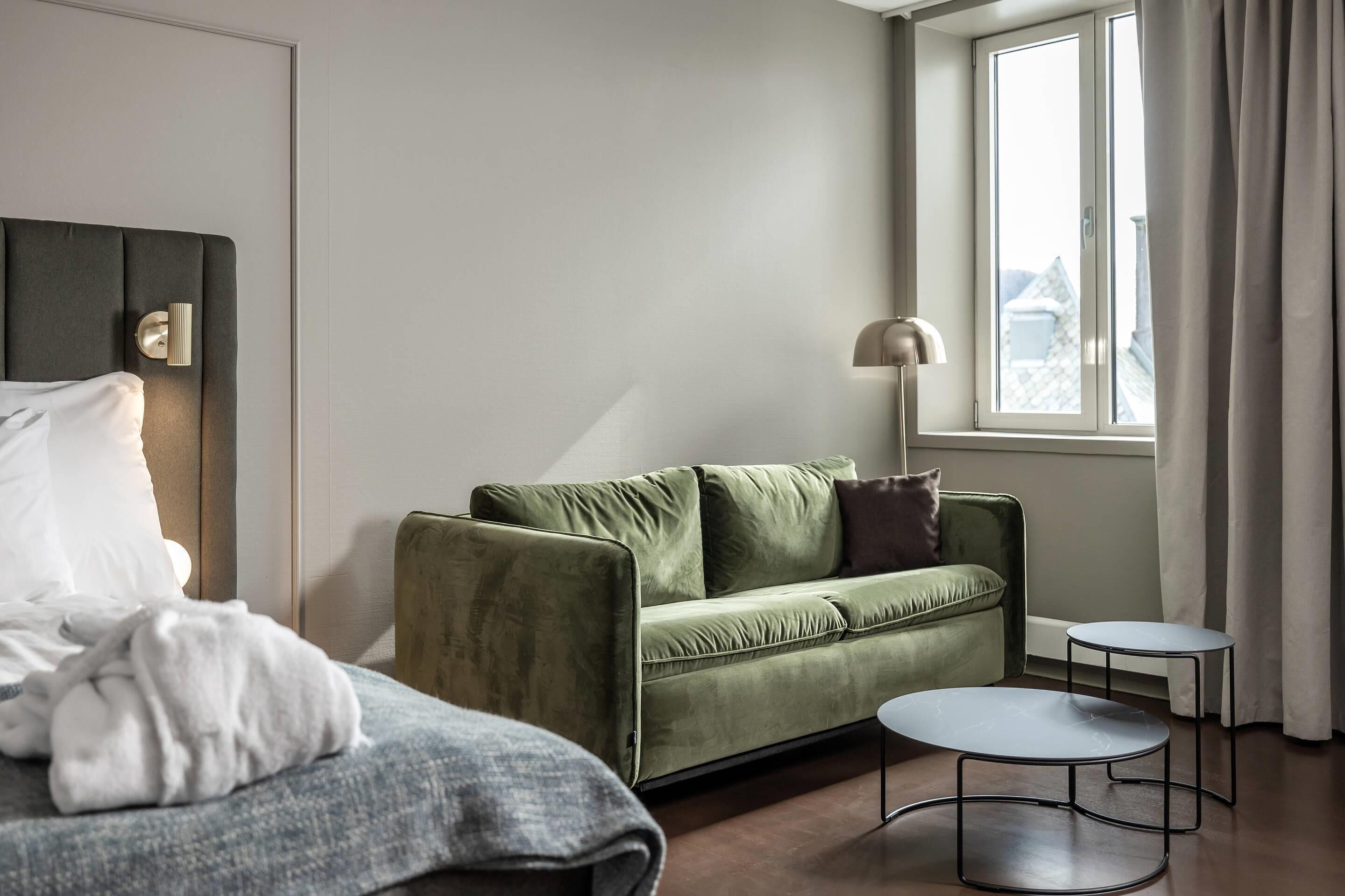 Une chambre d’hôtel moderne avec un canapé vert, une table ronde, un lit avec un oreiller blanc et une fenêtre avec des rideaux.