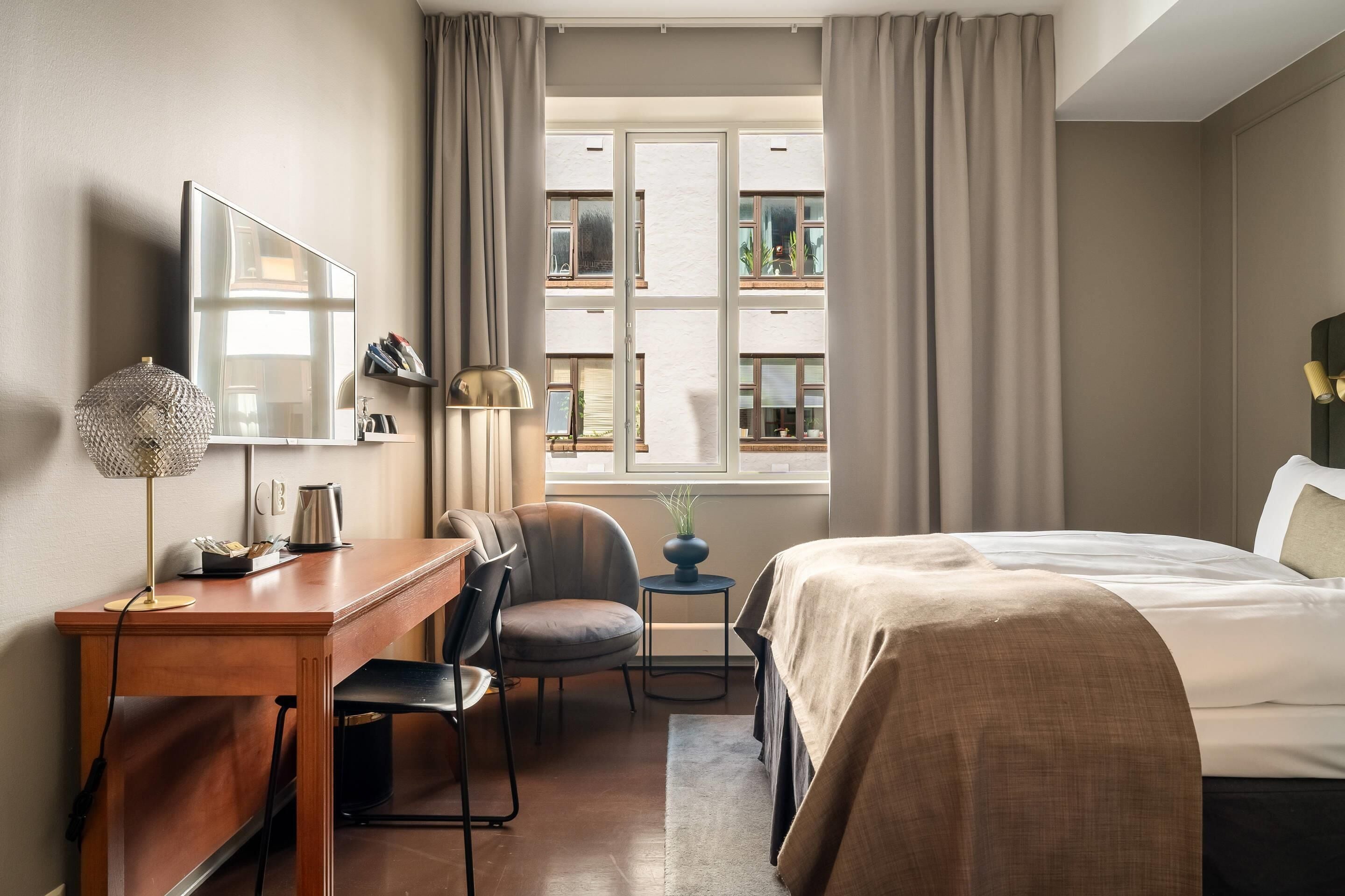 A hotel room with a bed, a desk with a chair, a small table with a lamp, and a window with curtains.