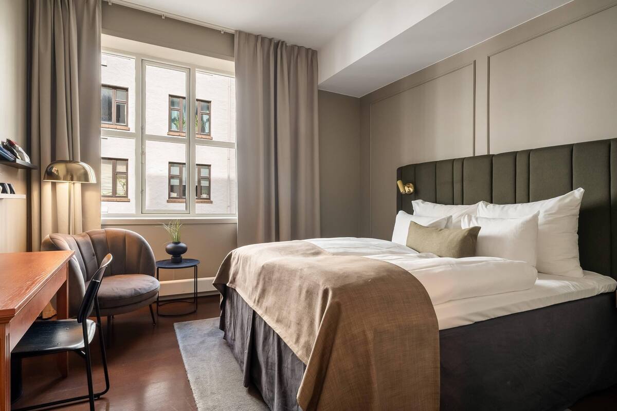 Ein Hotelzimmer mit Bett, Schreibtisch, Stuhl, einem kleinen Tisch mit Pflanze und einem Fenster mit Vorhängen.