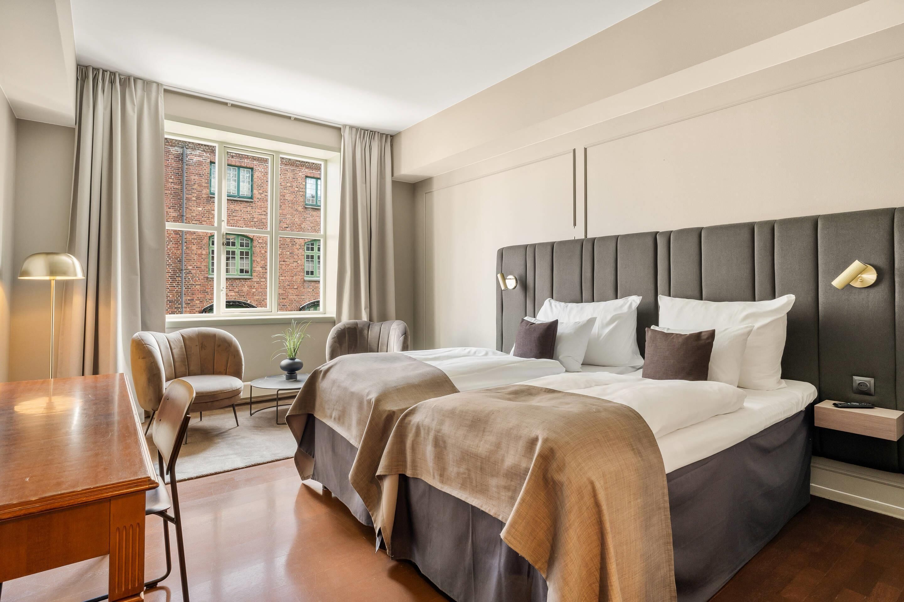 A modern hotel room with a large bed, a desk, and a chair. There is a window with curtains, a brick wall outside, and a nightstand with a lamp.