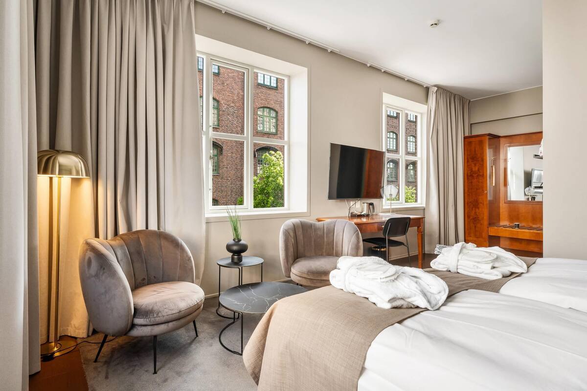 Ein Hotelzimmer mit einem Bett, zwei Sesseln, einem Schreibtisch, einem Fernseher und einem Fenster mit Vorhängen.