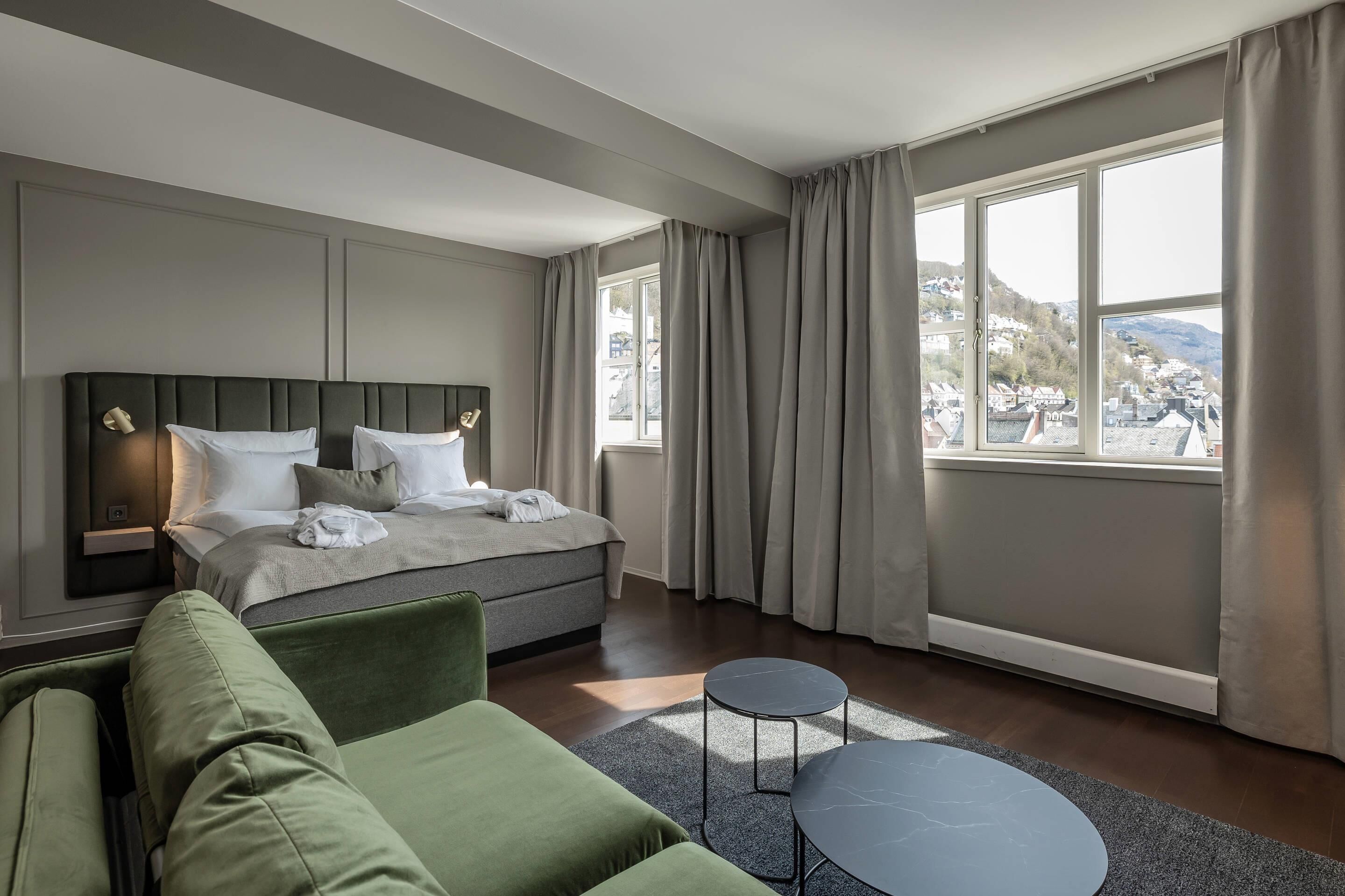 A modern hotel room with a large bed, a green sofa, two small tables, and a view of a town and hills through large windows.