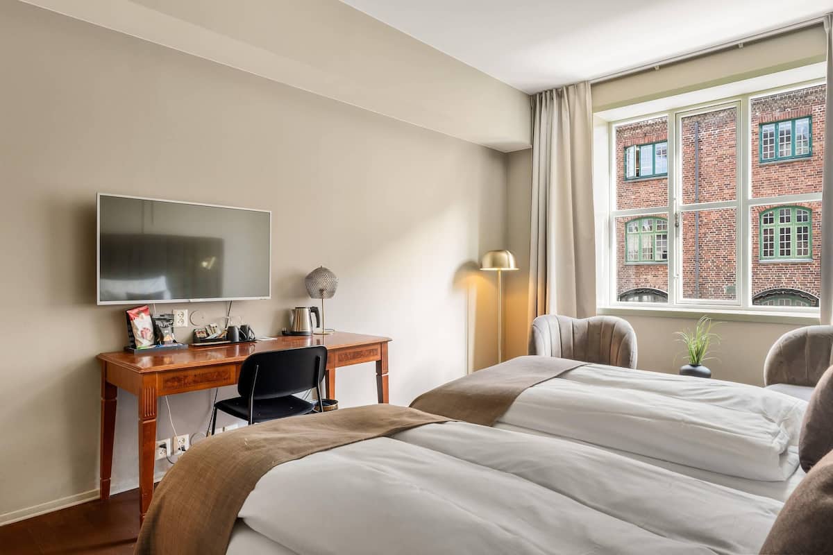 Ein Hotelzimmer mit einem Bett, einem Schreibtisch mit Fernseher, einer Lampe und Blick auf ein Backsteingebäude durch ein Fenster.