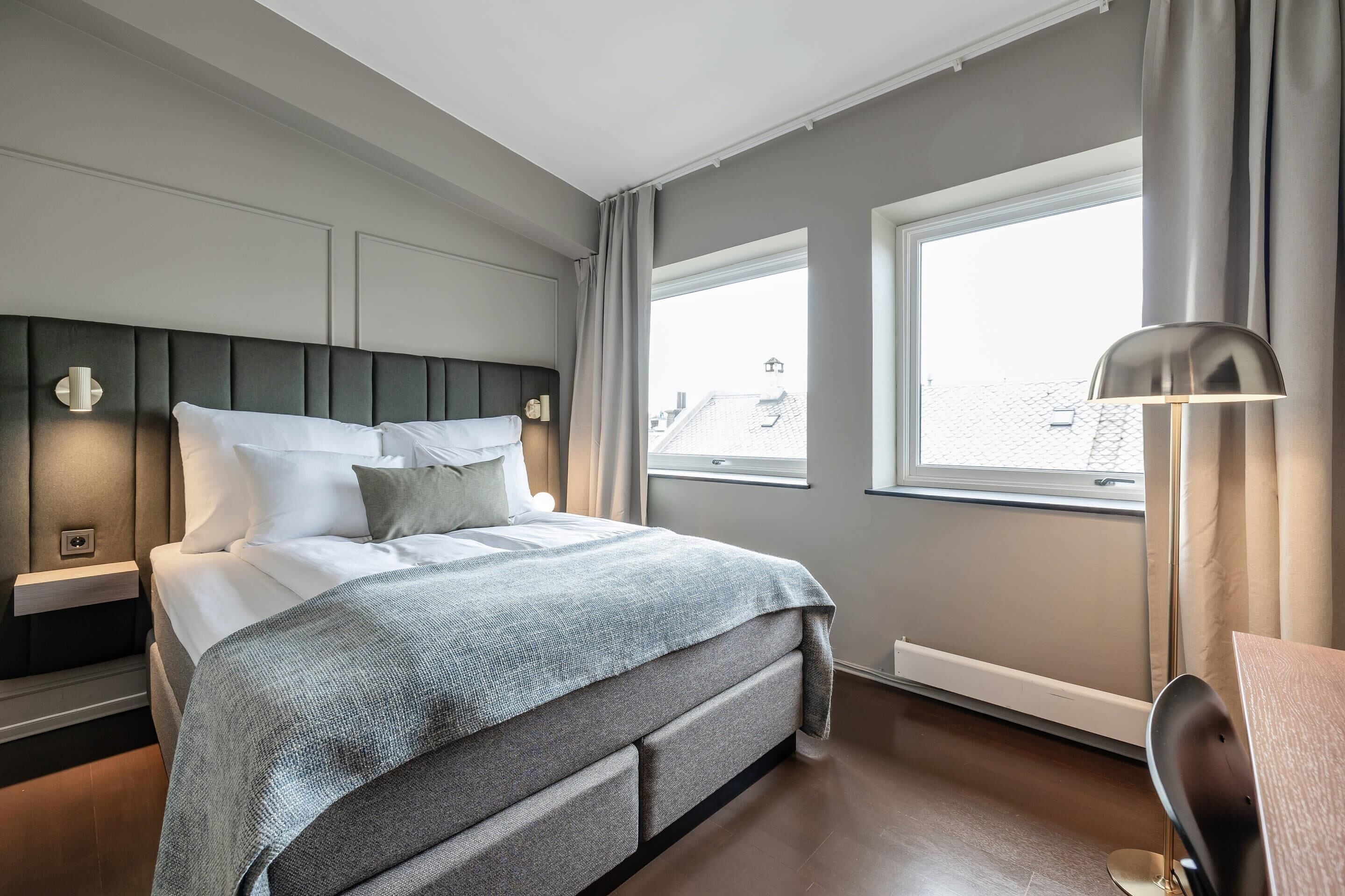 Una habitación de hotel moderna con una cama grande, una mesita de noche, una lámpara y una ventana con cortinas.