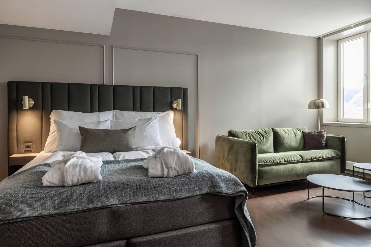 Ein modernes Hotelzimmer mit einem großen Bett, einem grünen Sofa, einem kleinen runden Tisch und einem Fenster mit Aussicht.