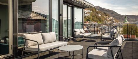 A modern outdoor seating area with sofas and chairs, large windows, and a view of a town and mountains.