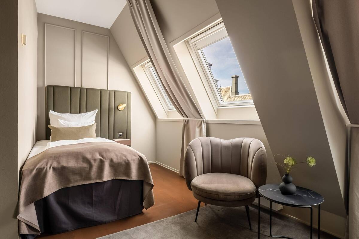 Ein Schlafzimmer mit einem Bett, einem Sessel, einem kleinen Tisch mit einer Vase und einem Fenster mit Vorhängen.