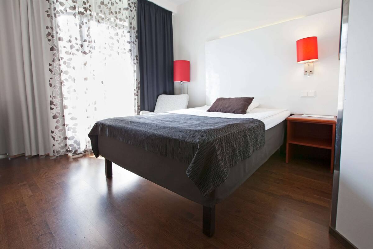 Ein Hotelzimmer mit einem Bett, einem Nachttisch, einer roten Lampe und einem Fenster mit Vorhängen.