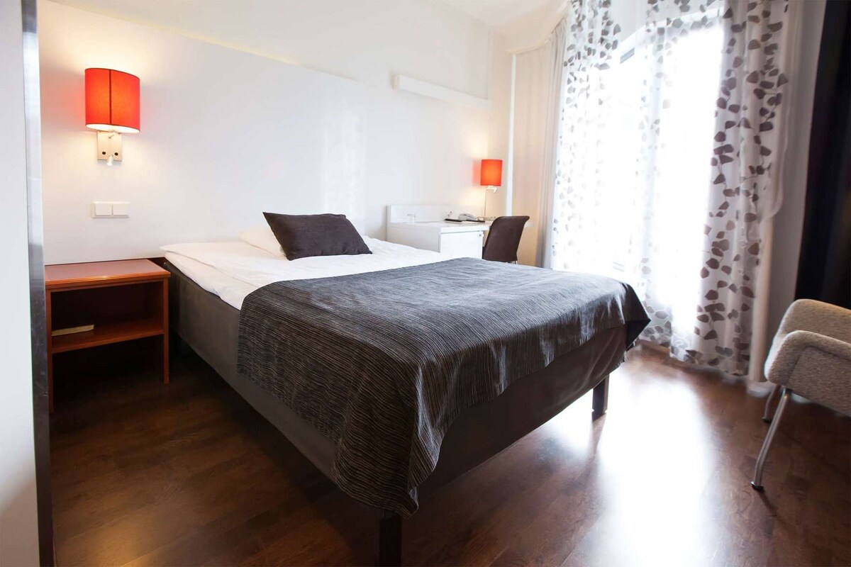 A hotel room with a bed, bedside tables, lamps, a chair, and a window with curtains.