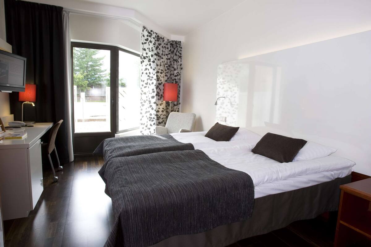 Ein Hotelzimmer mit zwei Betten, einem Schreibtisch, einem Stuhl, einem Fernseher und einem Fenster mit Vorhängen.