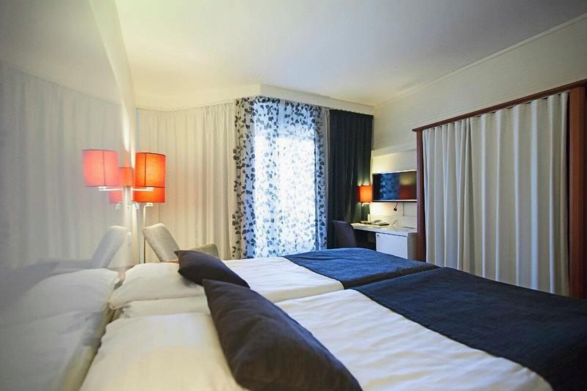 A hotel room with two beds, a desk, a chair, a lamp, and a window with curtains.