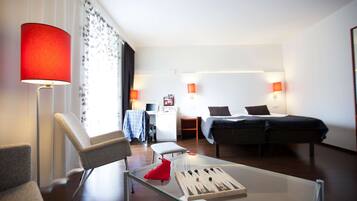A modern hotel room with a glass coffee table, a red lamp, a bed with a dark bedspread, a desk with a laptop, and a chair.
