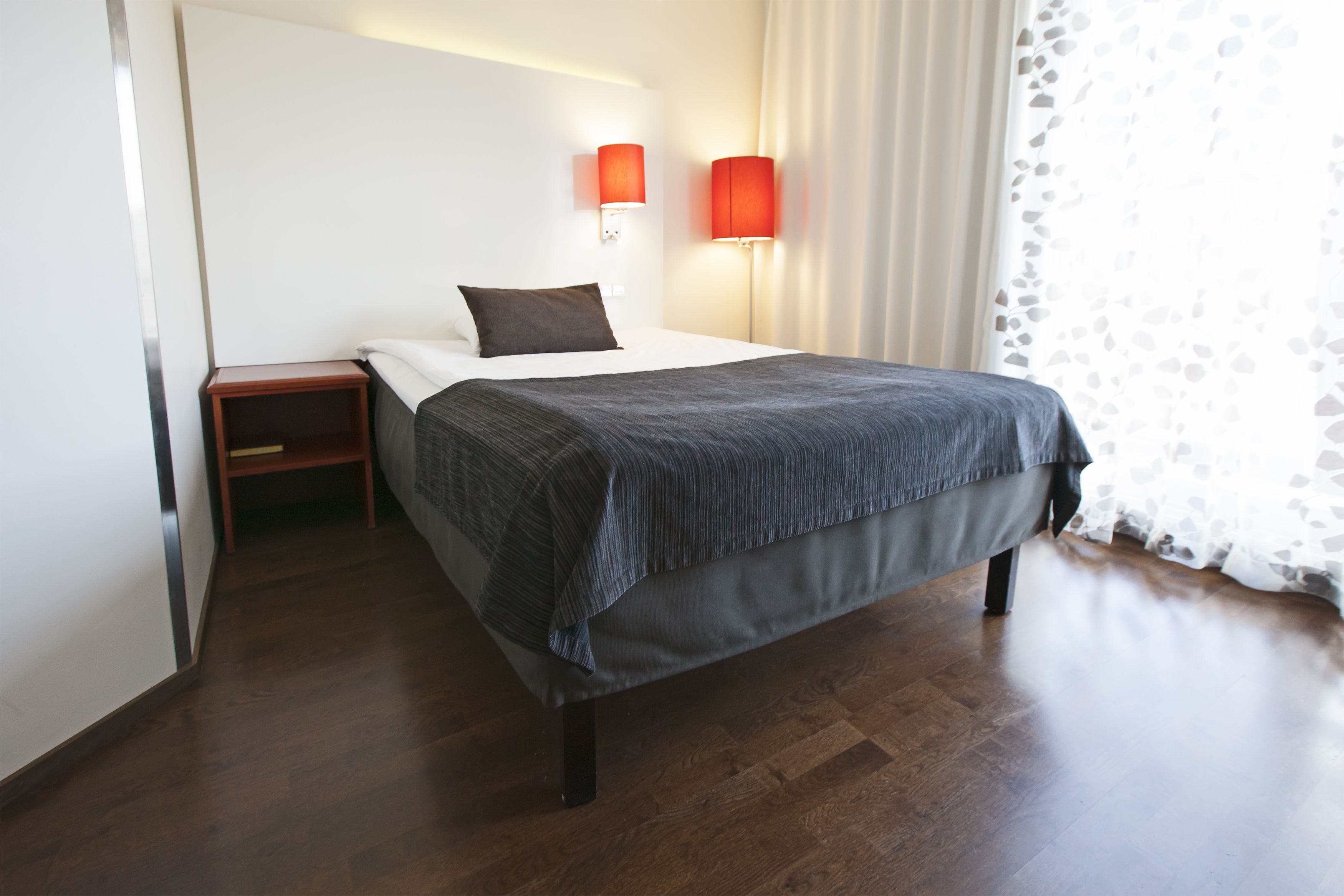 A neatly made bed with a grey blanket, a wooden nightstand, and two red wall lamps.