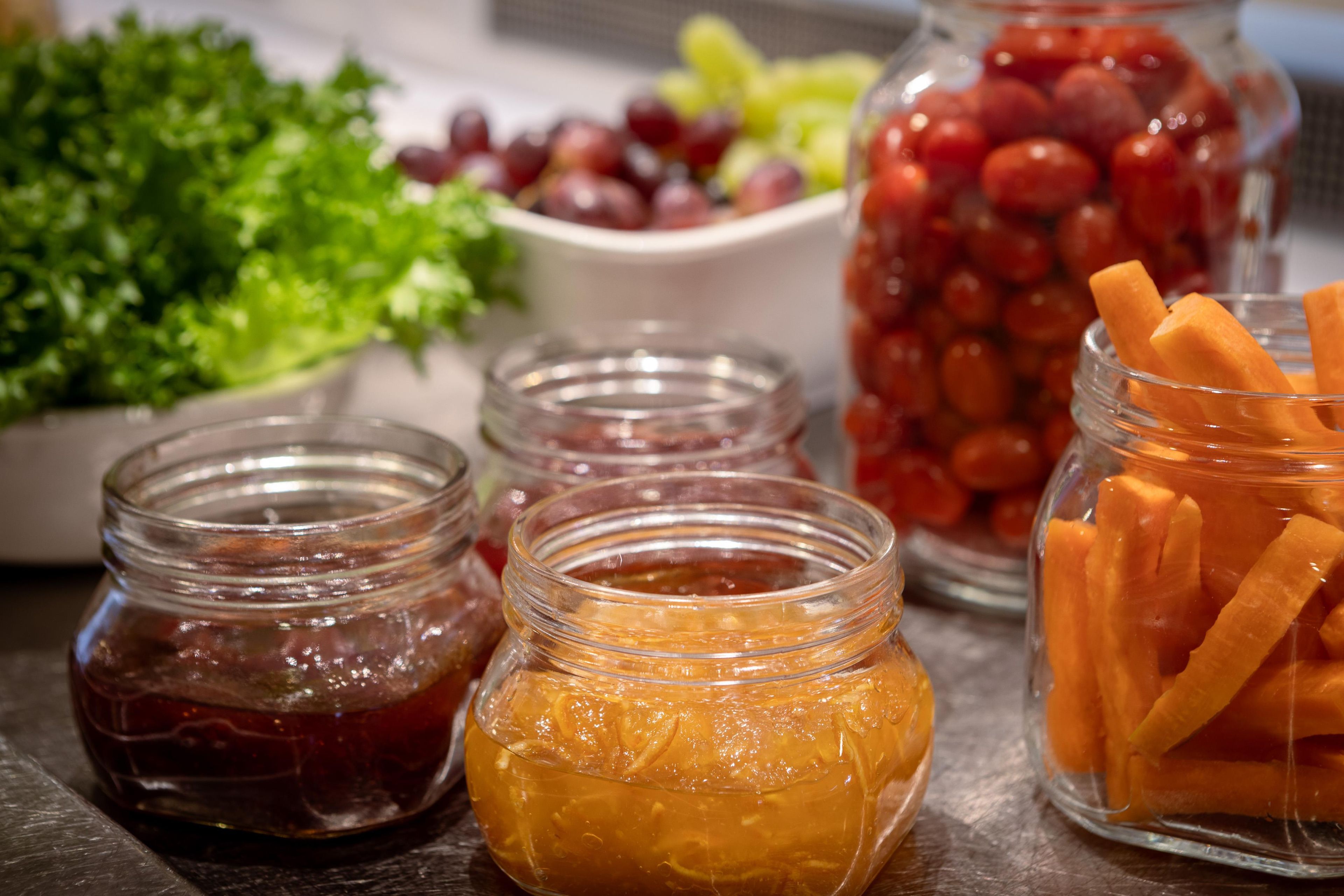 Uma mesa com diversos alimentos, incluindo potes de compotas, uma tigela de uvas e um recipiente com palitos de cenoura.