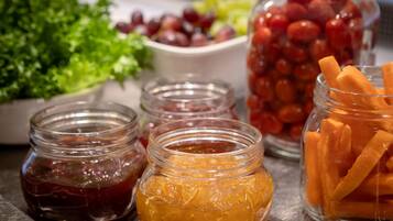 Uma mesa com diversos alimentos, incluindo potes de compotas, uma tigela de uvas e um recipiente com palitos de cenoura.