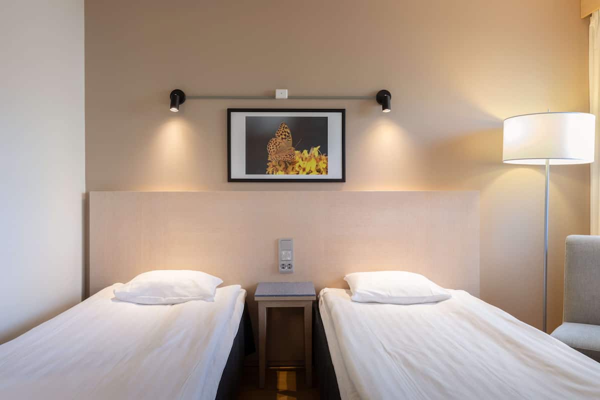 Hotellihuone, jossa on kaksi erillistä sänkyä, yöpöytä, lamppu ja seinällä oleva kehystetty kuva.