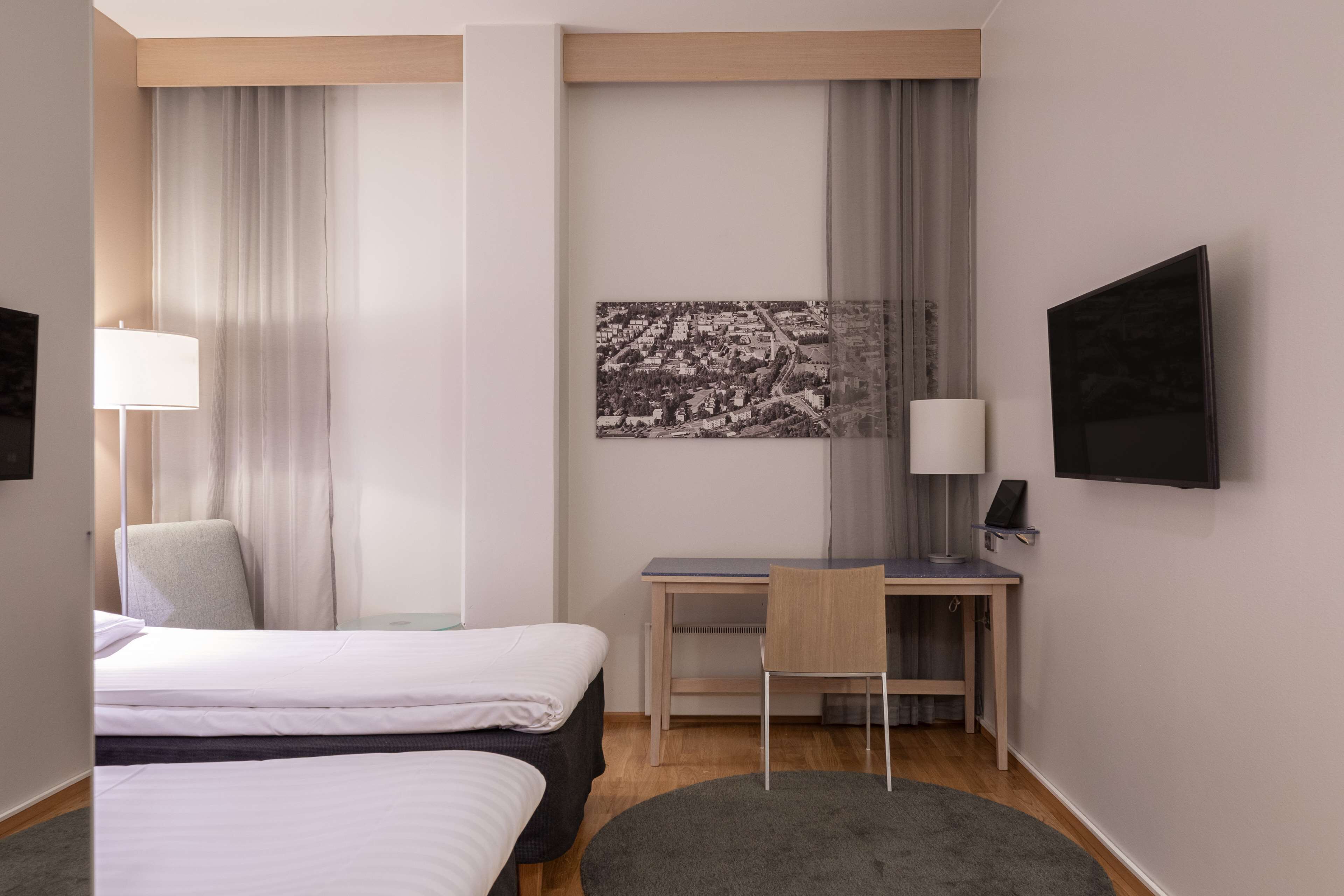 A hotel room with two beds, a desk, a chair, a TV, and a large cityscape picture on the wall.