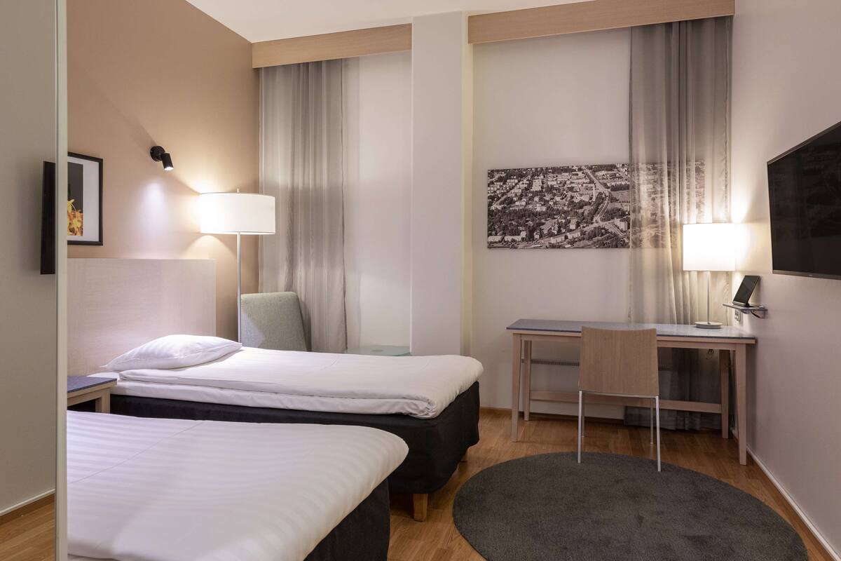 Hotellihuone, jossa on kaksi sänkyä, työpöytä, tuoli, televisio ja suuri ikkuna, jossa on verhot.