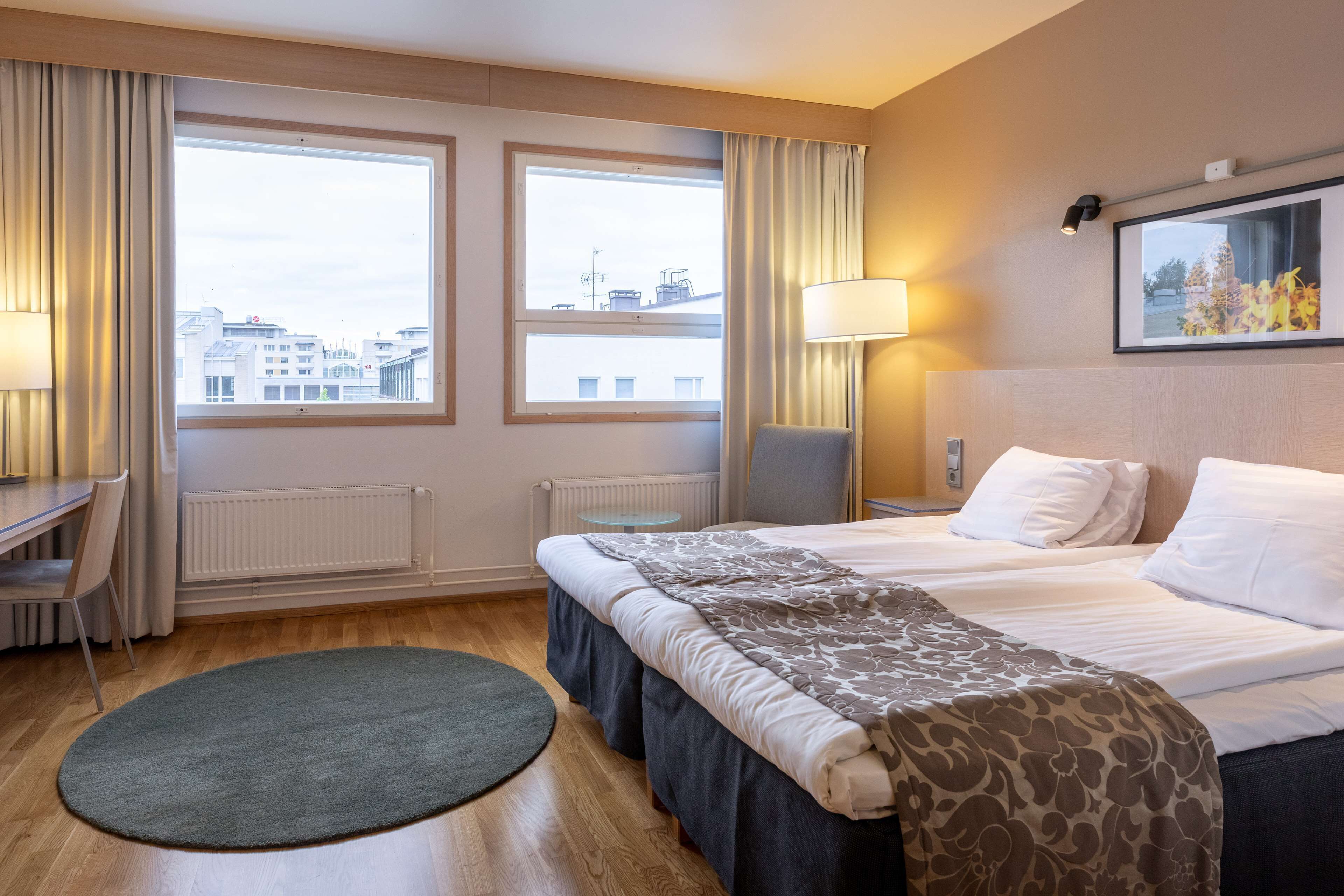 A hotel room with a bed, two windows, a desk, a chair, a lamp, and a view of the cityscape.