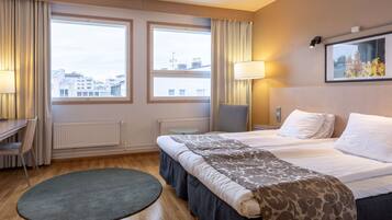 A hotel room with a bed, two windows, a desk, a chair, a lamp, and a view of the cityscape.