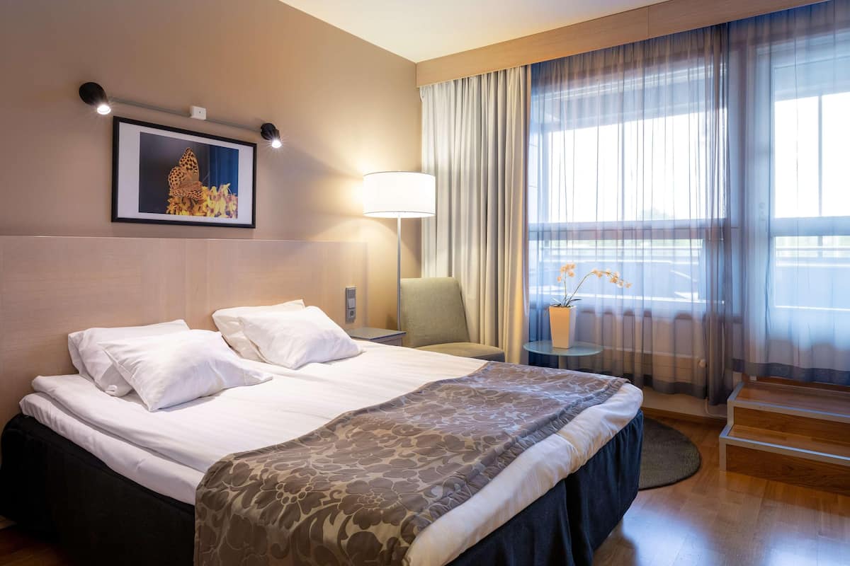 Hotellihuone, jossa on sänky, tuoli, lamppu, taulu seinällä ja ikkuna, jossa on verhot.