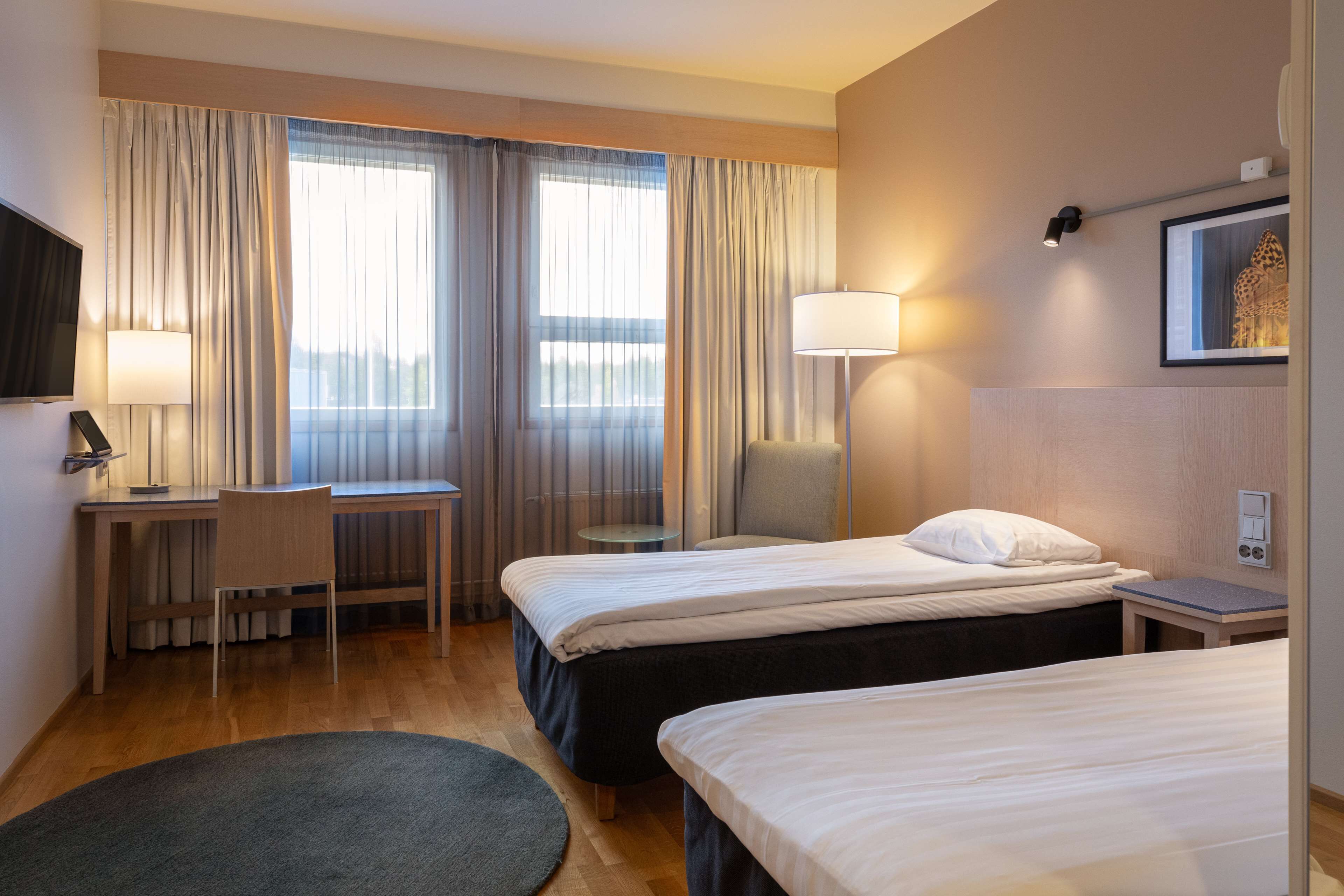 Habitación de hotel con dos camas, un escritorio, una silla, una lámpara y un cuadro en la pared.
