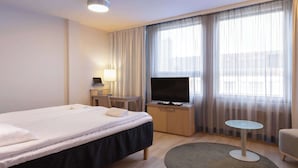 A hotel room with a bed, a desk with a TV, a small table, and a window with curtains.