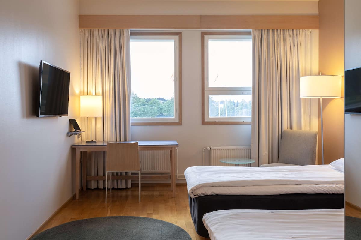 Hotellihuone, jossa on kaksi sänkyä, työpöytä, tuoli, televisio ja kaksi ikkunaa, joissa on verhot.