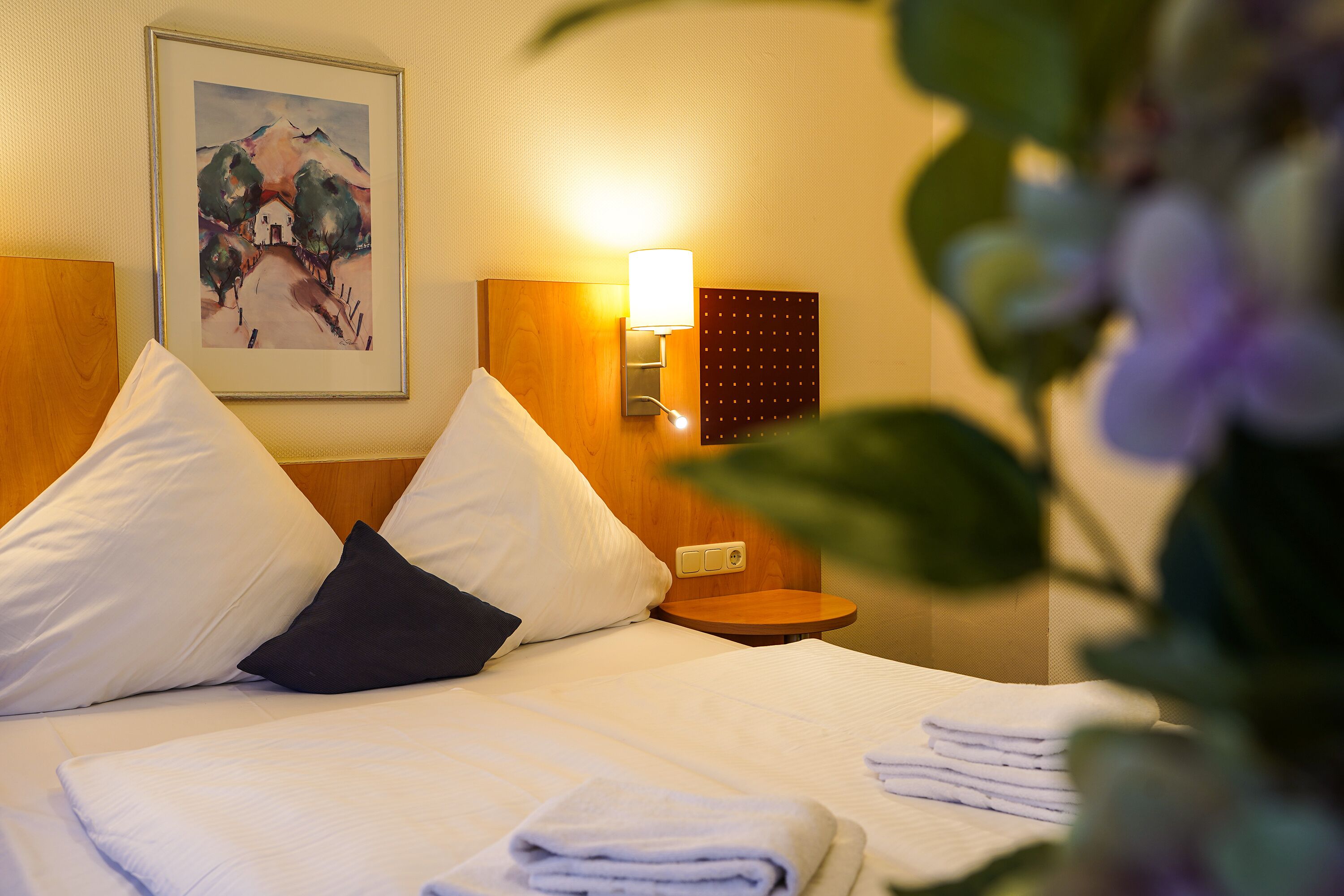 A neatly made bed with white and dark pillows, a wall-mounted lamp, and a framed painting above it.