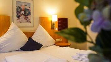 A neatly made bed with white and dark pillows, a wall-mounted lamp, and a framed painting above it.