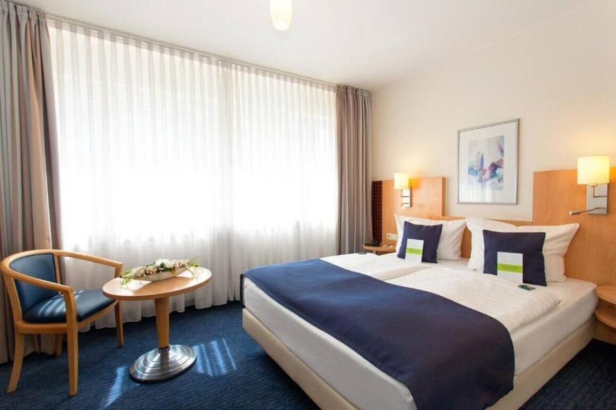 A hotel room with a large bed, two bedside tables with lamps, a chair, a small round table with a vase, and a large window with curtains.