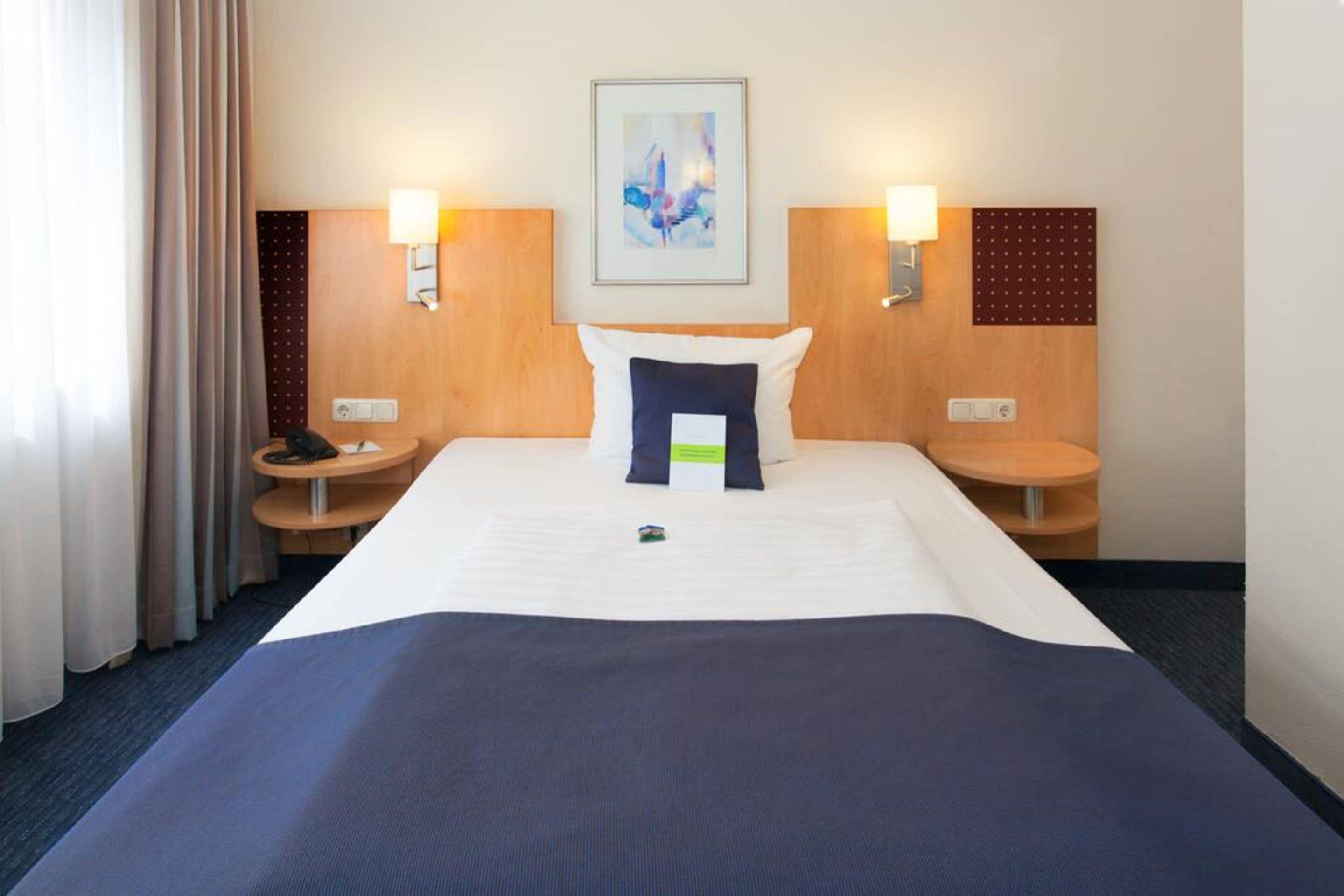 A hotel room with a large bed, two bedside tables, a headboard with a painting, and two wall-mounted lamps.