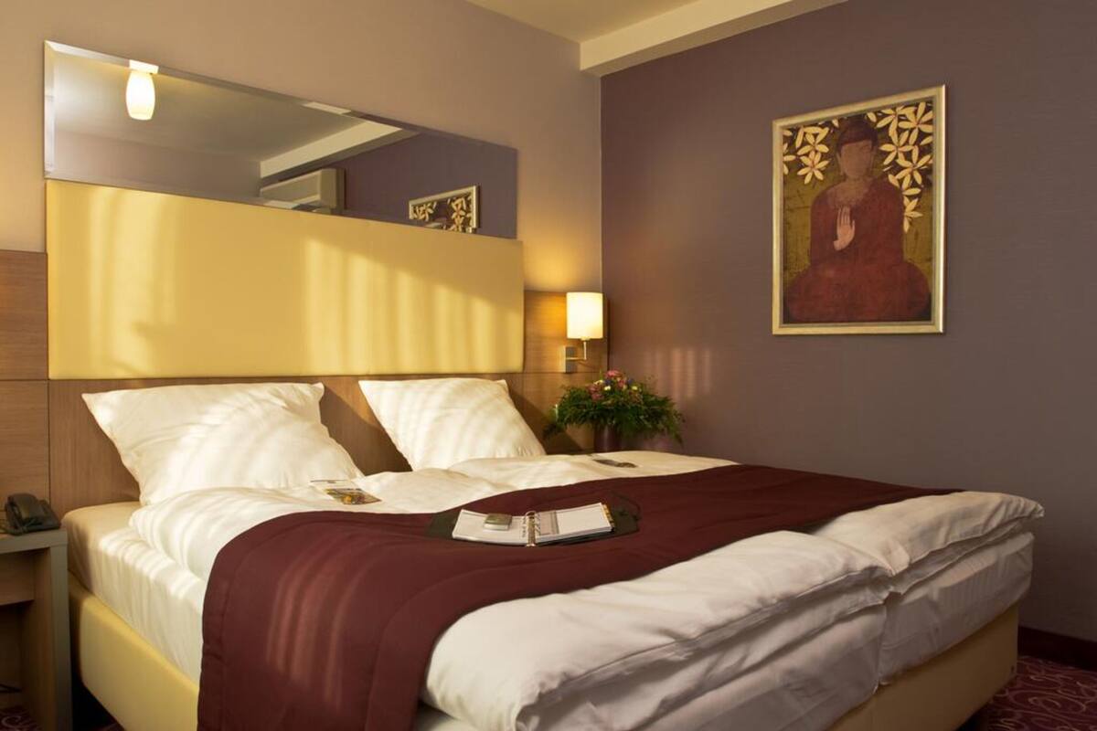 A hotel room with a bed, a bedside table with a lamp, a mirror above the bed, and a painting on the wall.