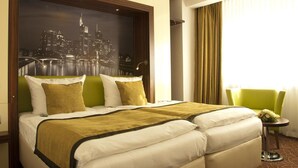 A hotel room with a large bed, two pillows, a green chair, a small table with a vase, and a cityscape view.
