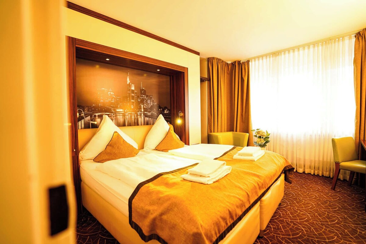 A hotel room with a large bed, two chairs, a window with curtains, and a cityscape view.