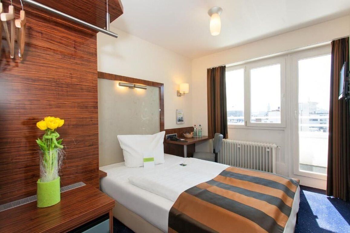 Habitación de hotel con cama, escritorio, silla, ventana con vistas y un jarrón con flores.