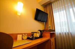 A hotel room with a flat-screen TV mounted on the wall, a desk with a laptop, a coffee maker, and a clock.