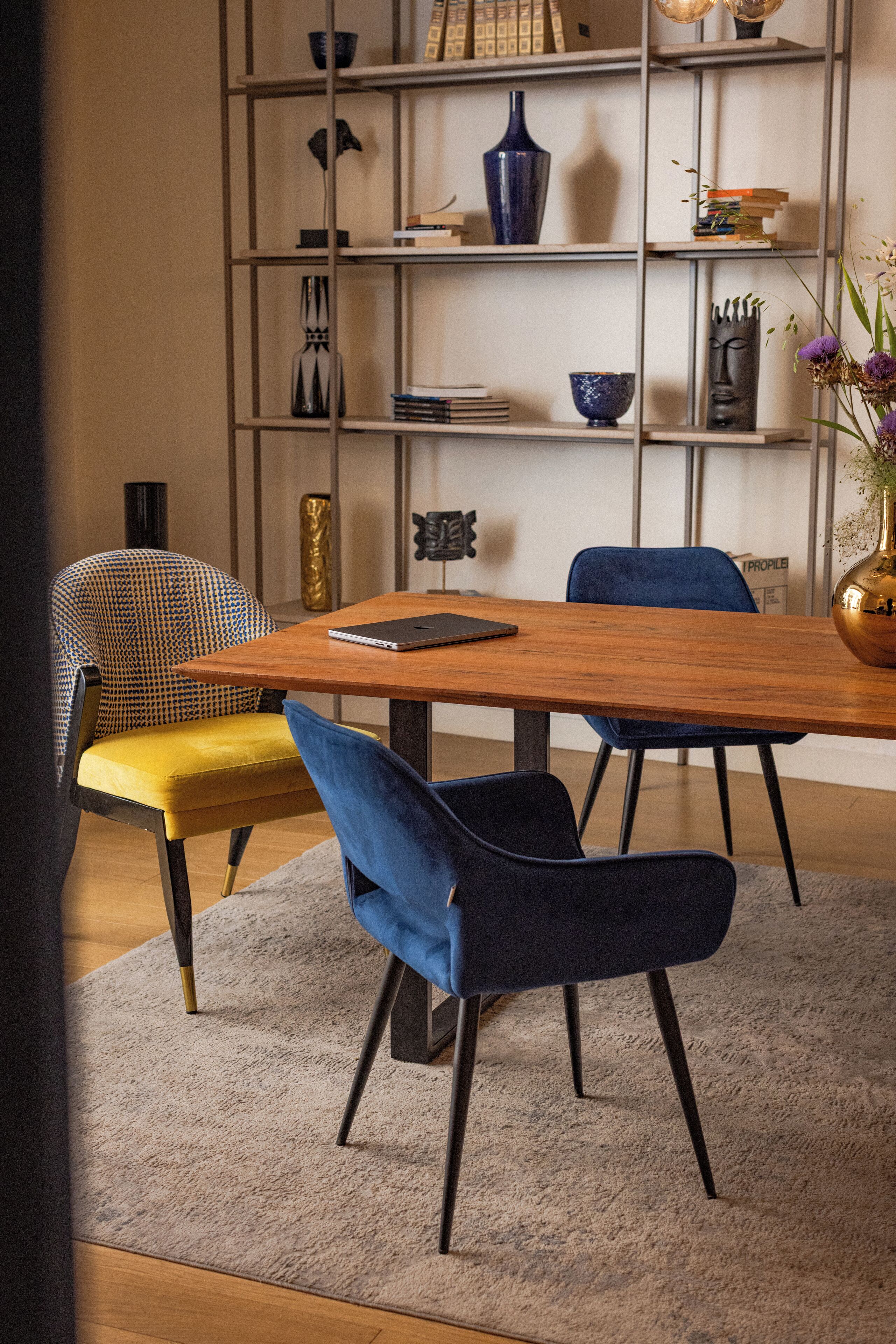 A dining area with a wooden table, blue and yellow chairs, a bookshelf with various decorative items, and a vase with flowers.
