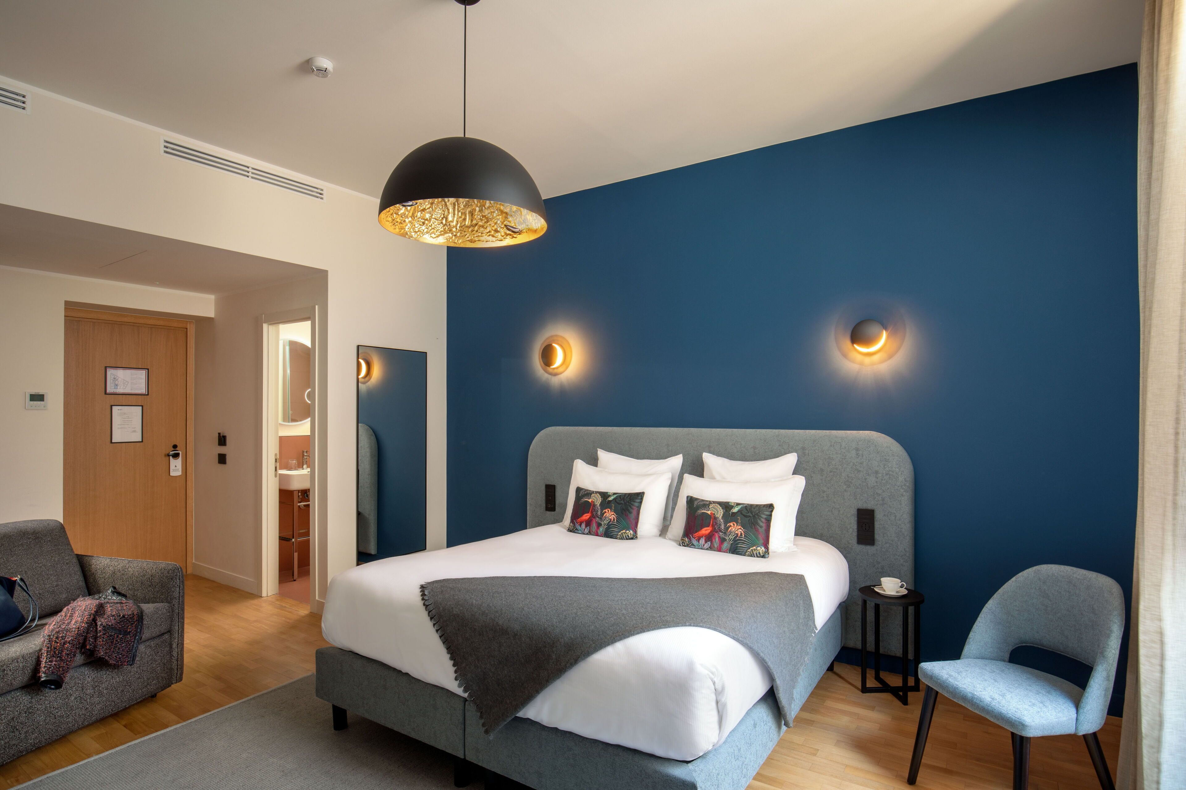 Ein modernes Hotelzimmer mit einem großen Bett, einem grauen Sessel, einem kleinen Beistelltisch und einer Deckenleuchte.