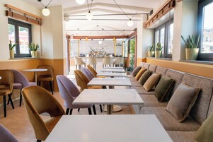A restaurant with a long counter, multiple tables, and chairs. There are potted plants on the windowsills and a bar area in the background.