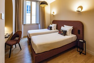 A hotel room with two beds, a chair, a small table, and a window with curtains.