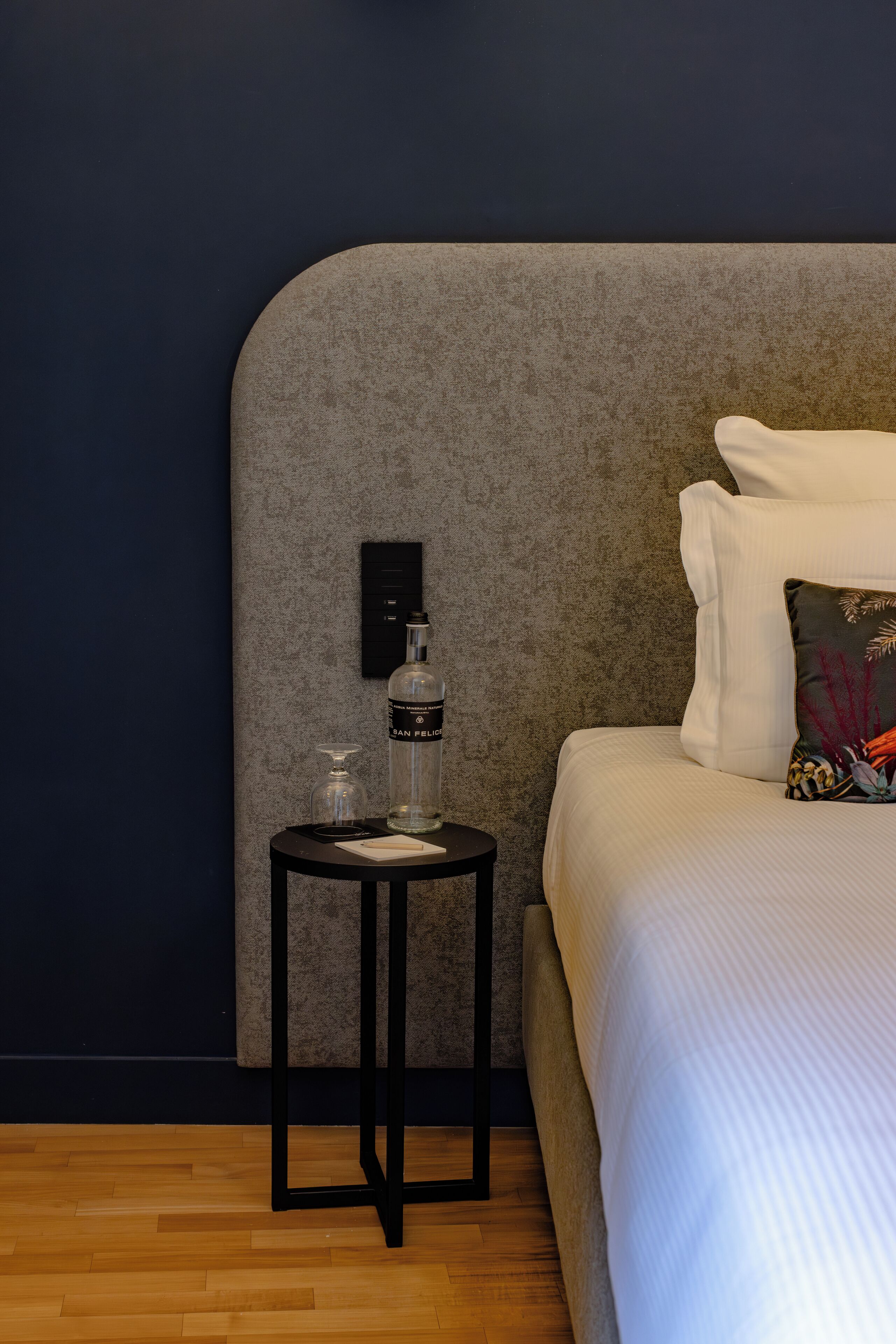 A neatly made bed with white linens, a small black side table with a bottle and a glass, and a wall-mounted control panel.